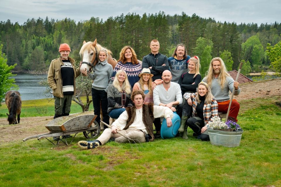 12 mennesker står sammen i en gruppe sammen med to hester, en gammel tretrillebår og et vaskevannsfat i sink brukt som blomsterpotte. Alle har på seg ullgensere